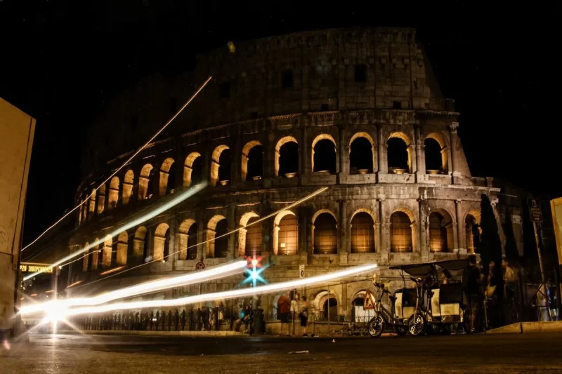 Coliseu iluminado à noite em Roma, com rastros de luz de veículos e pessoas.
