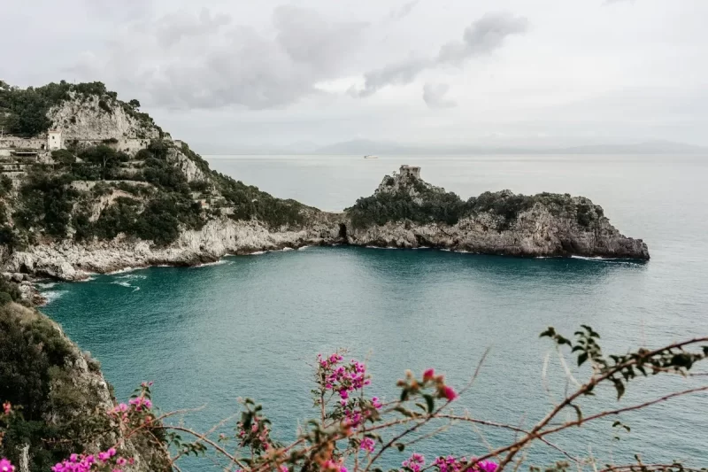 Vista panorâmica da Costa Amalfitana com mar turquesa, penhascos rochosos, ilhota com torre e flores roxas em primeiro plano.