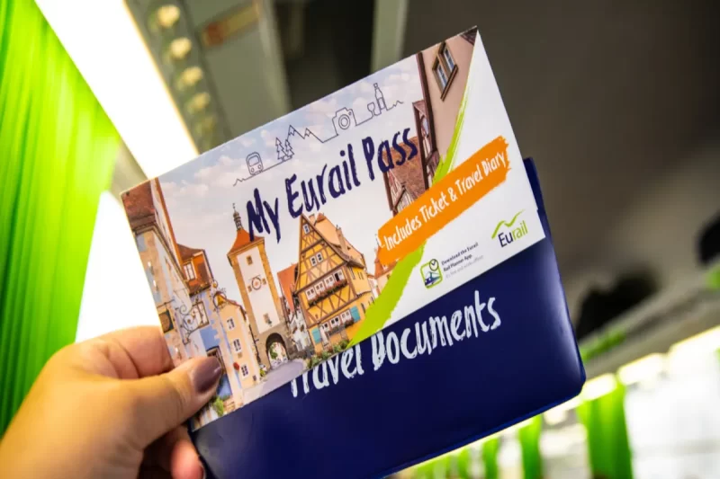 Mão segurando um Eurail Pass e diário de viagem com ilustrações de destinos europeus, promovendo a mobilidade de trem.