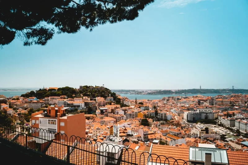 Vista aérea de Lisboa com telhados laranjas, Castelo de São Jorge e Rio Tejo ao fundo, enquadrada por árvore.