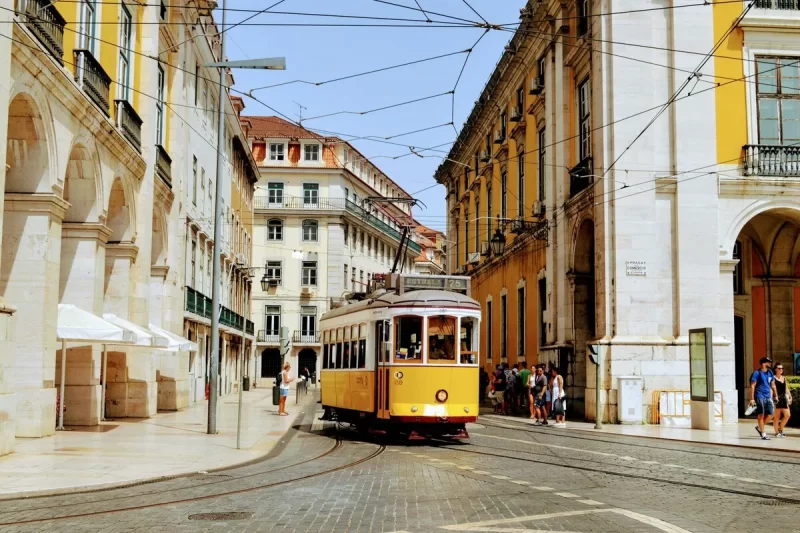 Elétrico amarelo clássico de Lisboa (bonde 28) em rua de paralelepípedos com edifícios históricos e pedestres.