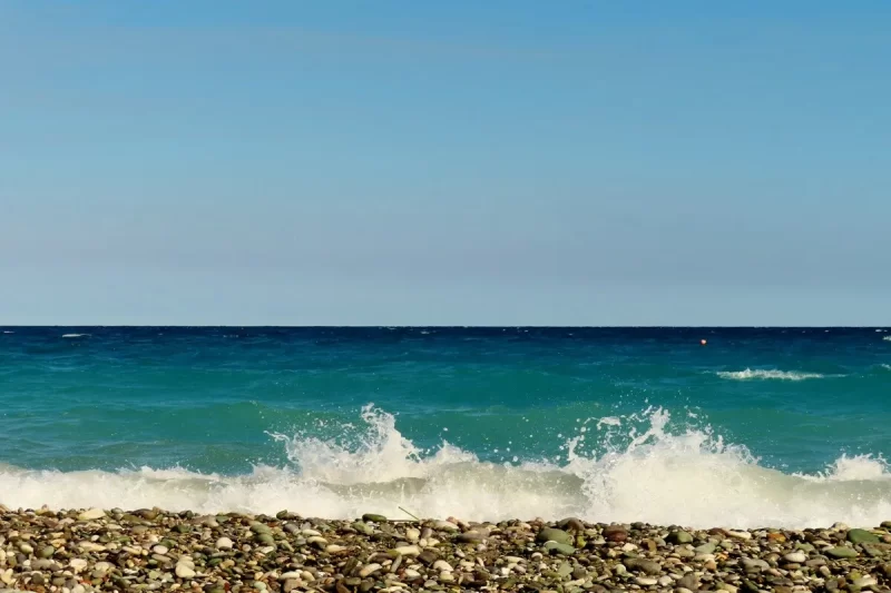 Ondas quebrando suavemente em uma praia de seixos coloridos sob um céu azul claro.