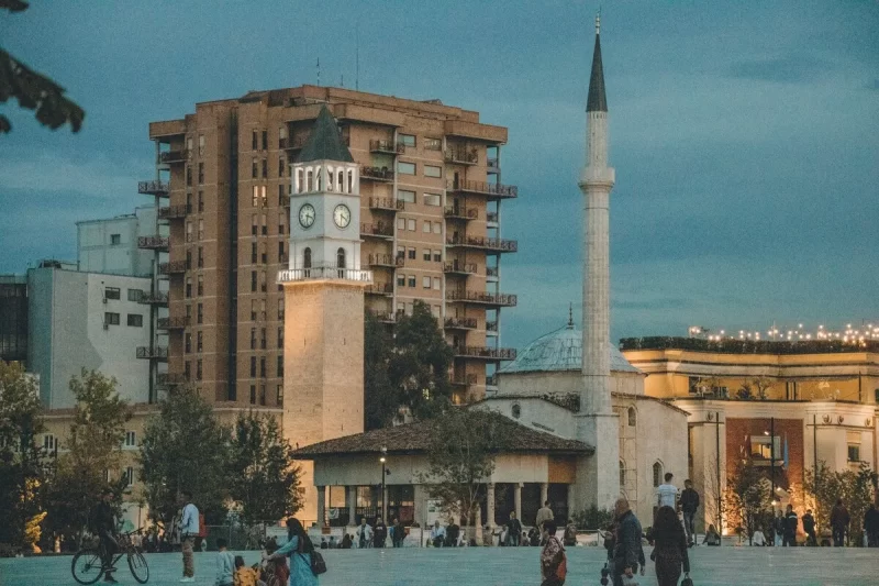 Vista ao entardecer da Praça Skanderbeg em Tirana, Albânia, com a Mesquita Et'hem Bey e a Torre do Relógio no centro, e pessoas passeando.