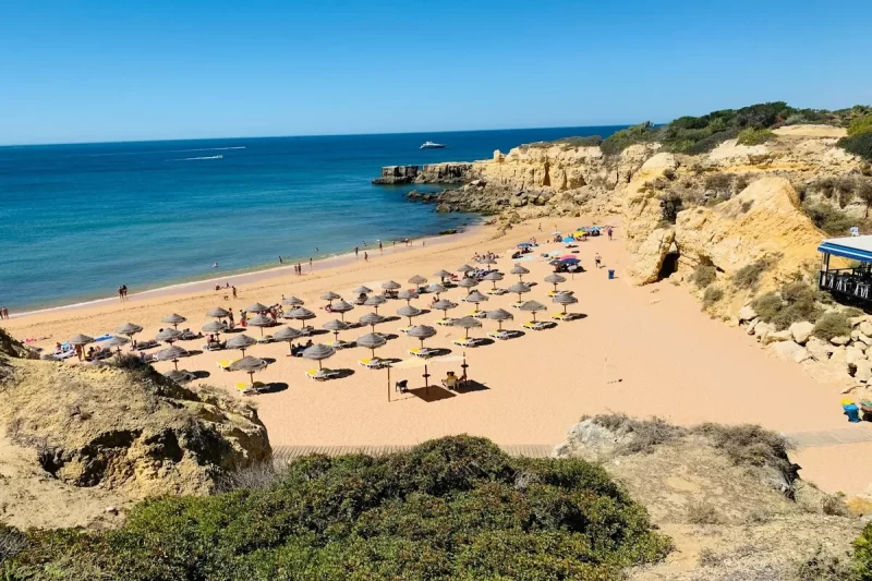 Praia ensolarada no Algarve com muitos guarda-sóis de palha, águas azuis, falésias douradas e vegetação costeira, barcos no horizonte.