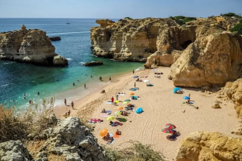 Vista aérea de uma praia no Algarve com falésias rochosas, águas claras, pessoas na areia com guarda-sóis, barcos no horizonte.