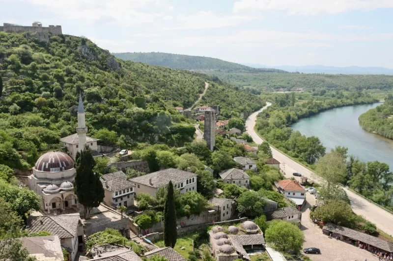 Vista panorâmica de uma cidade antiga nos Balcãs, com telhados de pedra, uma mesquita com cúpula e minarete, e um rio serpenteando por um vale verdejante sob um céu claro.