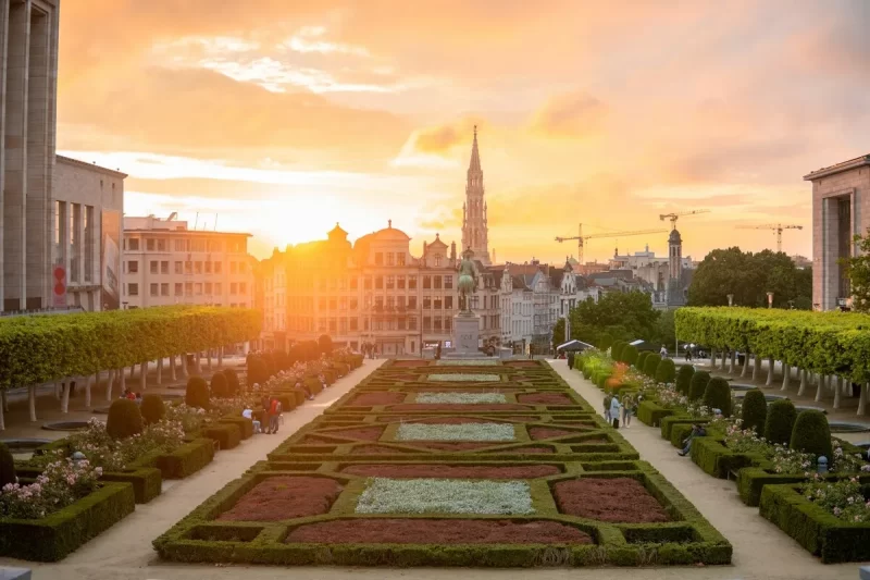 O icônico jardim do Mont des Arts em Bruxelas ao pôr do sol, com seus canteiros geométricos, arbustos podados e uma vista panorâmica da cidade e sua arquitetura, incluindo a torre do centro.