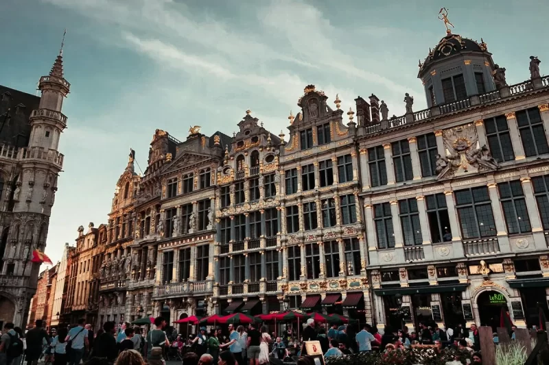 Vista de uma multidão na Grand Place de Bruxelas, com os edifícios guildistas ornamentados e dourados, e a torre da prefeitura ao lado esquerdo, sob um céu claro.