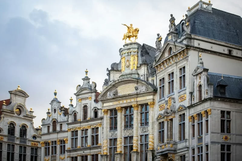 Edifícios históricos ornamentados com detalhes dourados na Grand Place em Bruxelas, apresentando arquitetura barroca e estátuas no telhado sob um céu nublado.