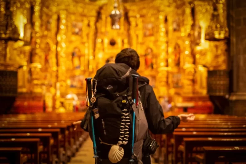 Peregrino com mochila e concha do Caminho de Santiago de costas, contemplando altar dourado de igreja, fim da jornada, momento de fé e gratidão.