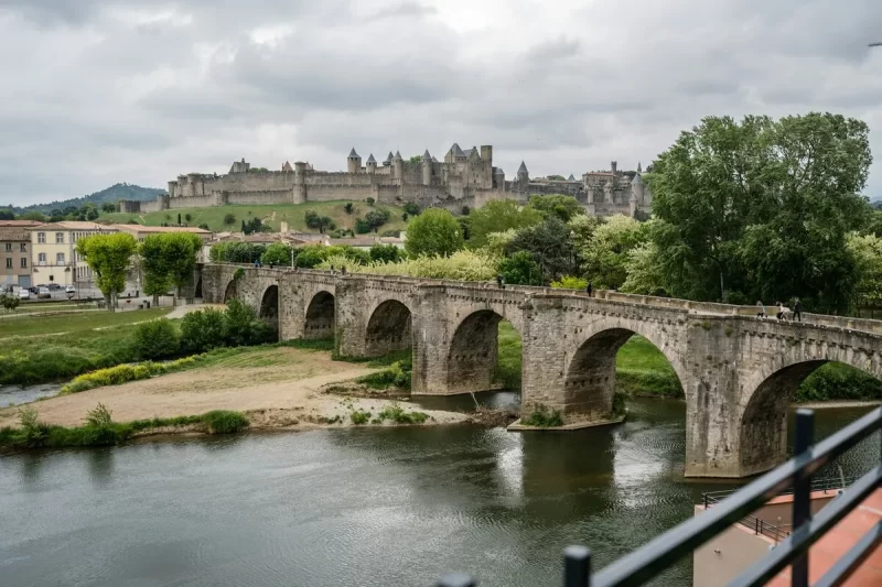 Vista panorâmica da cidade fortificada de Carcassonne ao fundo, com a Ponte Velha de arcos de pedra atravessando o rio Aude em primeiro plano, sob um céu nublado.