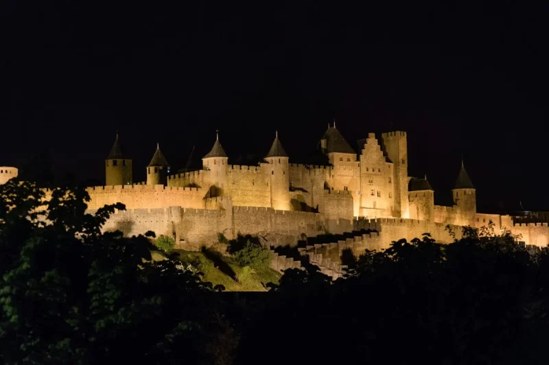 Vista noturna da cidade medieval de Carcassonne, com suas muralhas e torres iluminadas em dourado contra o céu escuro.