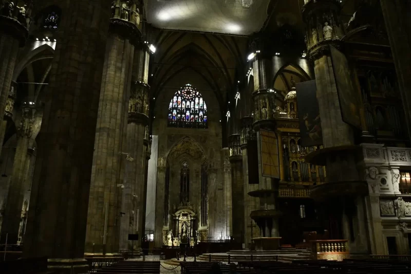 Interior escuro e imponente da Catedral de Milão (Duomo) com vitrais coloridos, altares ornamentados, colunas maciças e a grandiosidade da arquitetura gótica.