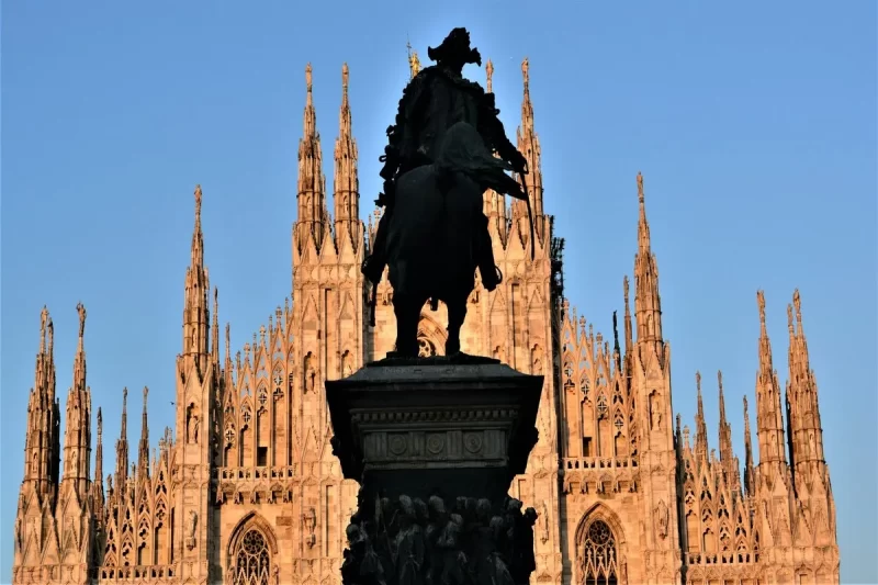 Silhueta escura de uma estátua equéstre em primeiro plano, com a Catedral de Milão (Duomo) iluminada ao fundo sob um céu azul, destacando seus pináculos góticos.