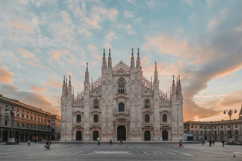 Vista frontal da grandiosa Catedral de Milão (Duomo) com seus pináculos góticos intrincados sob um céu vibrante de tons pastel ao entardecer, com poucas pessoas na praça.
