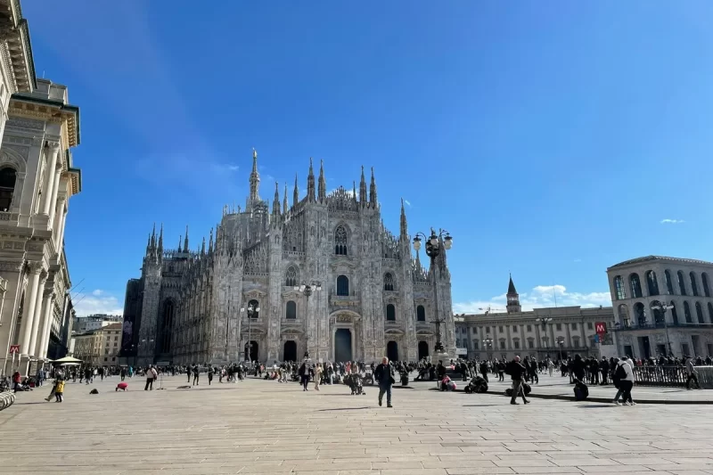 Vista ampla da majestosa Catedral de Milão (Duomo) com sua arquitetura gótica imponente, pináculos e estátuas, em uma praça movimentada sob um céu azul.