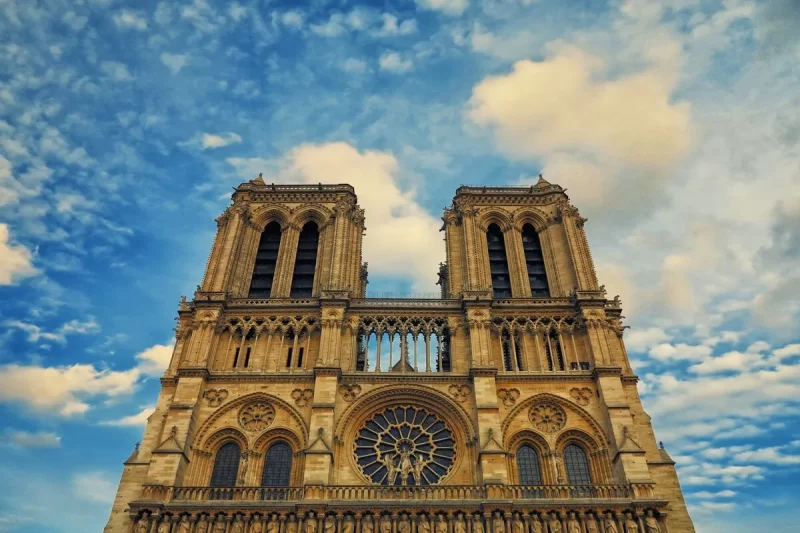 Vista frontal detalhada da Catedral de Notre-Dame em Paris, com suas duas torres, a grande rosácea central e esculturas góticas sob um céu azul com nuvens.