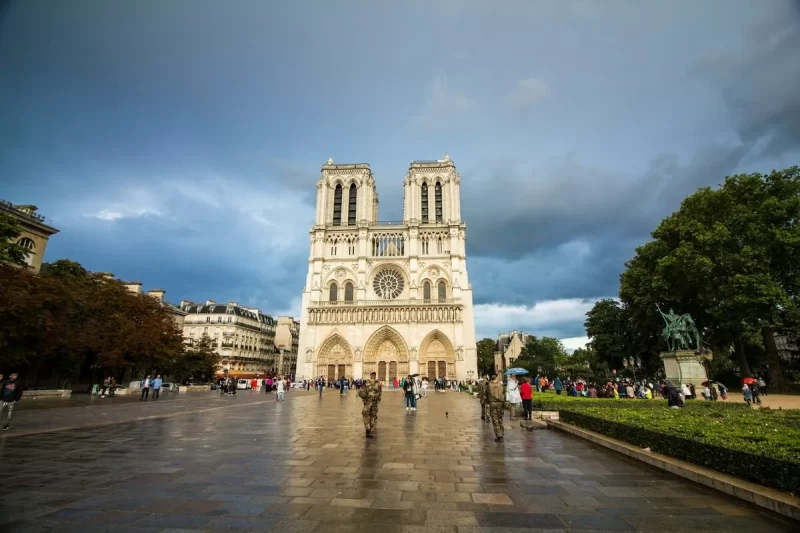 Vista ampla da Catedral de Notre-Dame em Paris, com suas torres e fachada imponentes, em frente a uma praça movimentada sob um céu dramático com nuvens escuras.