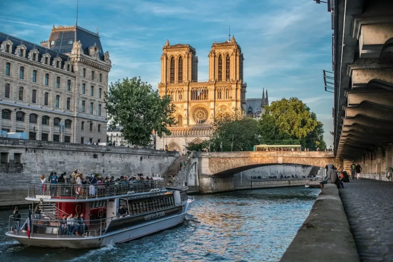 Vista externa da Catedral de Notre-Dame em Paris, com suas torres góticas, às margens do Rio Sena, onde um barco turístico navega.