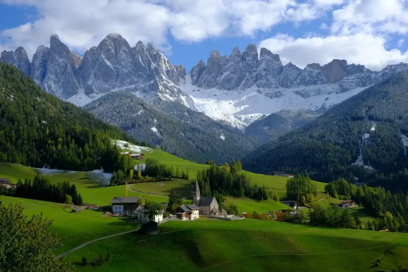 Vista panorâmica de uma pitoresca aldeia no vale de Val di Funes nas Dolomitas, com campos verdes, casas tradicionais e picos nevados das montanhas Geisler ao fundo.