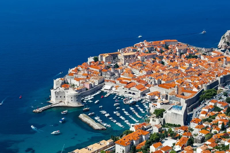 Vista aérea da cidade murada de Dubrovnik, Croácia, com telhados de cerâmica alaranjada, muros de pedra e pequenos barcos ancorados no mar azul