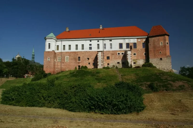 Um castelo histórico de tijolos e telhado vermelho em uma colina verde sob um céu azul, representando a arquitetura característica da Europa Central.