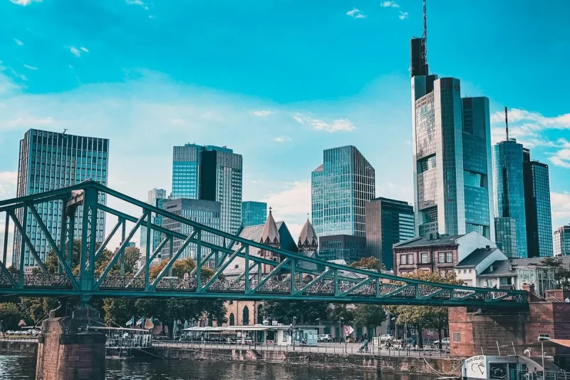 Skyline moderno de Frankfurt, Alemanha, com arranha-céus imponentes, uma ponte de ferro verde em primeiro plano e o rio Main.