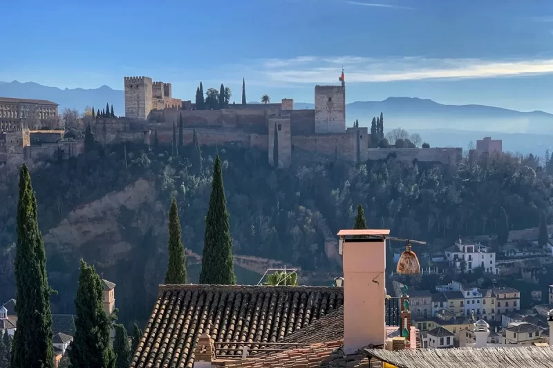 Vista panorâmica de Granada com o complexo da Alhambra no alto de uma colina, torres históricas e telhados de terracota em primeiro plano, ressaltando a beleza de Granada.