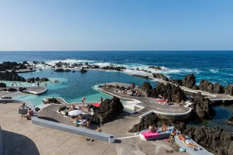 Piscinas naturais de rocha vulcânica em Porto Moniz, Ilha da Madeira, com pessoas nadando e relaxando sob o sol e o oceano Atlântico ao fundo.