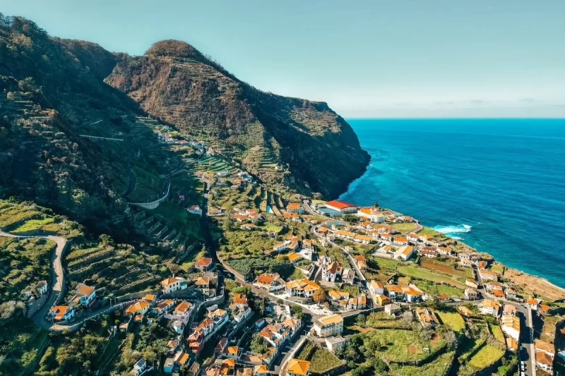 Vista aérea panorâmica de uma vila costeira na Ilha da Madeira, com casas coloridas e telhados de terracota espalhadas pelas encostas verdes das montanhas íngremes que se encontram com o azul vibrante do oceano Atlântico.