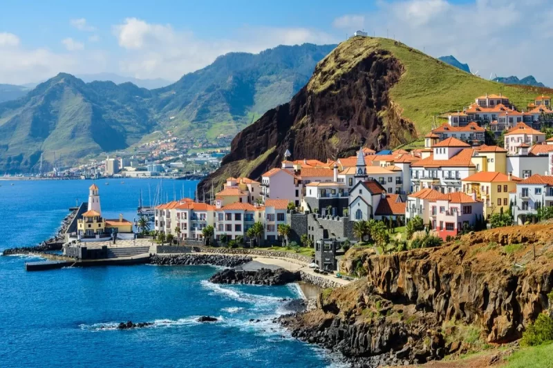 Panorama costeiro da Ilha da Madeira, com uma vila de casas coloridas e telhados de terracota, um farol e montanhas ao fundo, refletindo a beleza de um dos destinos mais procurados da Ilha da Madeira.