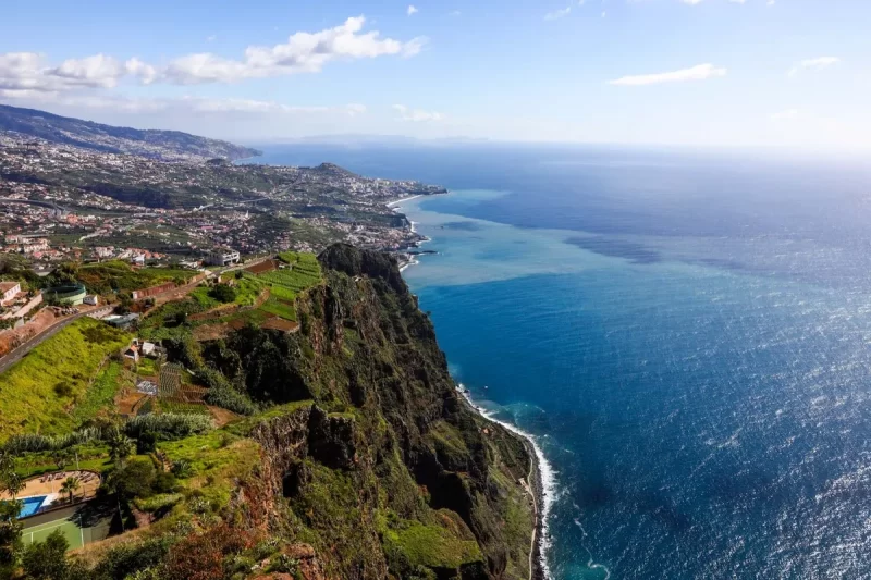 Vista aérea deslumbrante da costa da Ilha da Madeira, com falésias verdes, casas e cidades costeiras se estendendo até o oceano Atlântico azul.