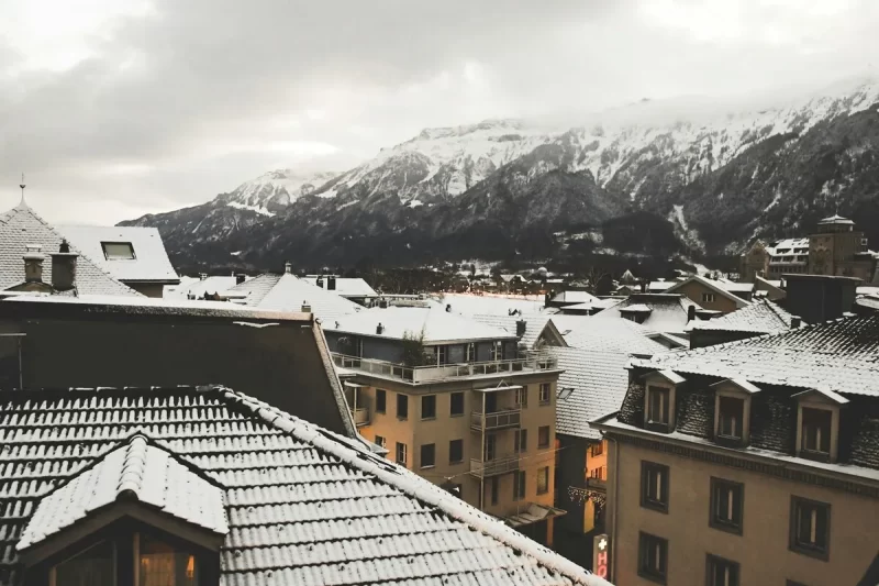 Vista aérea de telhados cobertos de neve na cidade de Interlaken, Suíça, com montanhas nevadas e encobertas por nuvens ao fundo, sob um céu nublado de inverno.
