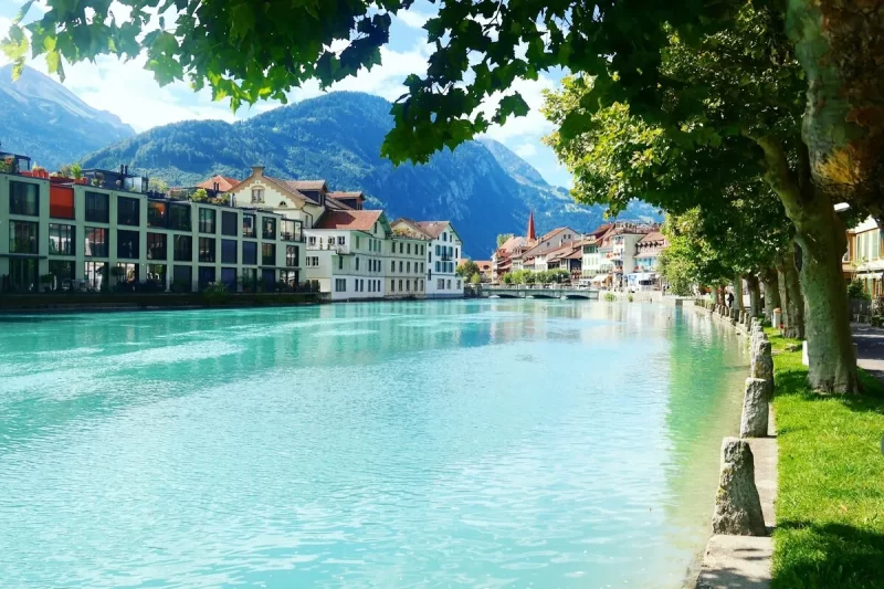 O Rio Aare de cor turquesa brilhante fluindo através da cidade de Interlaken, Suíça, com edifícios charmosos, pontes e montanhas verdes ao fundo.