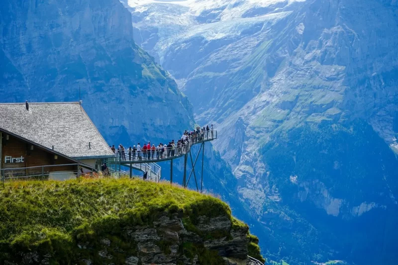 Pessoas em uma passarela de metal suspensa (Cliff Walk) no Grindelwald First, Suíça, com vistas deslumbrantes das montanhas nevadas e vales azuis ao redor de Interlaken.