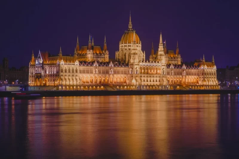 O Parlamento Húngaro em Budapeste, iluminado à noite, refletindo suas luzes douradas nas águas escuras do Rio Danúbio, sob um céu azul-arroxeado.