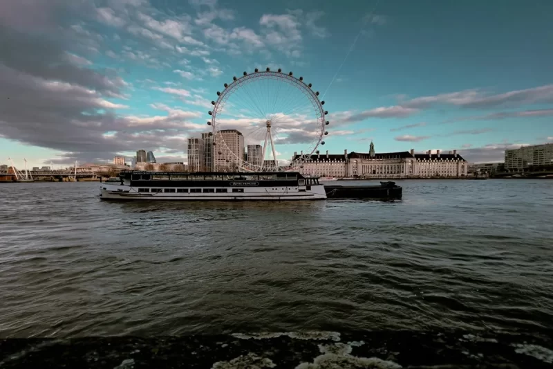 A London Eye e a paisagem urbana de Londres ao entardecer, com o Rio Tâmisa em primeiro plano e um barco de passeio navegando. O céu apresenta tons de azul escuro e nuvens.