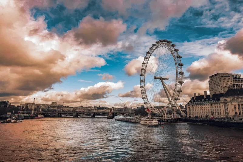A London Eye vista do Rio Tâmisa em um dia com céu parcialmente nublado, com edifícios da cidade e pontes visíveis ao fundo, e barcos atracados próximos à estrutura da roda gigante.