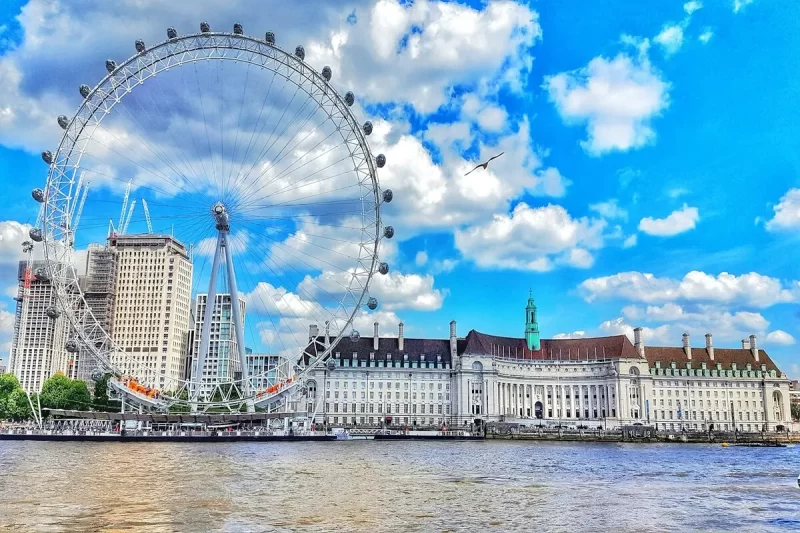 A London Eye, a grande roda gigante, sob um céu azul vibrante com nuvens brancas, às margens do Rio Tâmisa, com edifícios históricos e modernos ao fundo em Londres.