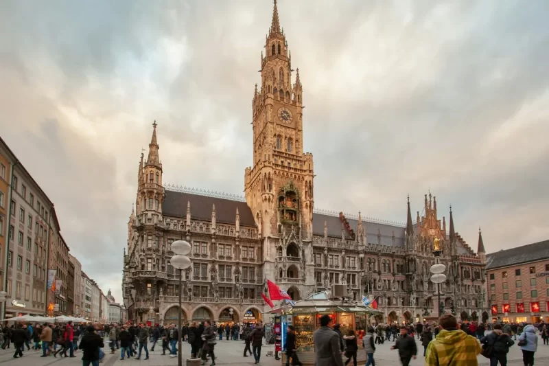Marienplatz em Munique, Alemanha, com a Nova Prefeitura (Neues Rathaus) e sua torre gótica, repleta de pessoas e lojas.