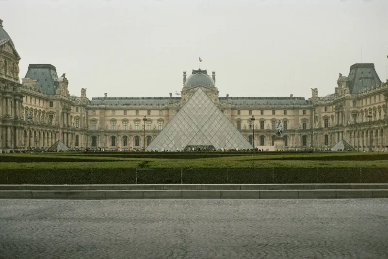 A Pirâmide de vidro do Museu do Louvre em Paris, França, com o majestoso palácio histórico ao fundo sob um céu nublado, vista de frente.