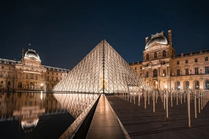 Pirâmide de vidro do Museu do Louvre iluminada à noite, refletindo-se em um espelho d'água em frente à arquitetura clássica do palácio sob um céu escuro.