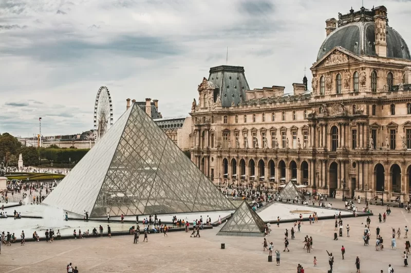 Vista panorâmica do Museu do Louvre em Paris, França, com a pirâmide de vidro principal, as edificações históricas do palácio e muitos visitantes na praça, sob um céu parcialmente nublado.