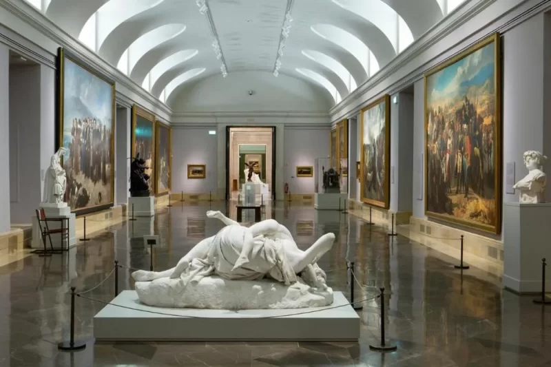 Longa galeria do Museu do Prado com grandes pinturas emolduradas e esculturas clássicas em exposição, incluindo uma proeminente escultura de mármore no centro do salão.