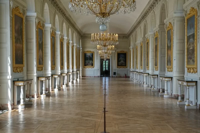 Interior grandioso do Palácio de Versalhes com lustres dourados, colunas brancas, pinturas ornamentadas e piso de madeira polido, refletindo a opulência histórica.