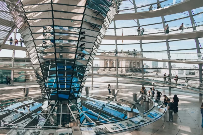 Interior da cúpula de vidro do Reichstag em Berlim, com a impressionante estrutura em espiral de espelhos e rampas, e visitantes caminhando.