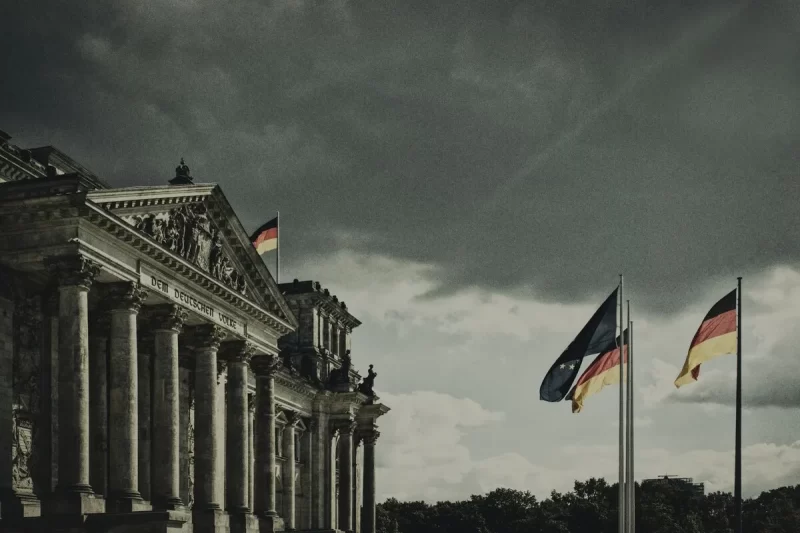 O imponente edifício do Reichstag em Berlim sob um céu dramático e parcialmente nublado, com bandeiras alemãs e outras bandeiras estaduais tremulando em frente à fachada.