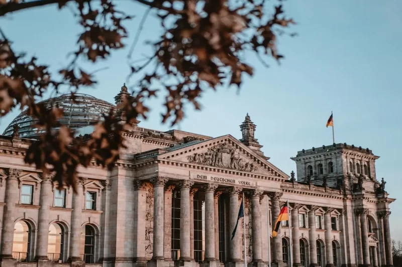 O edifício do Reichstag em Berlim, com sua cúpula de vidro e fachada neoclássica, em um dia ensolarado com um céu claro e galhos de árvore em primeiro plano com folhas secas.