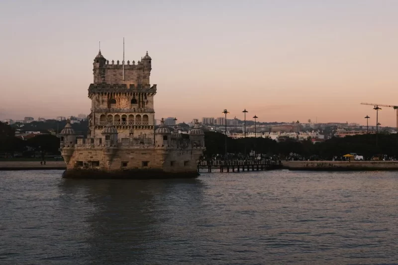 A icônica Torre de Belém em Lisboa ao pôr do sol, com suas fortificações iluminadas em tons quentes e a cidade ao fundo, destacando sua beleza histórica.