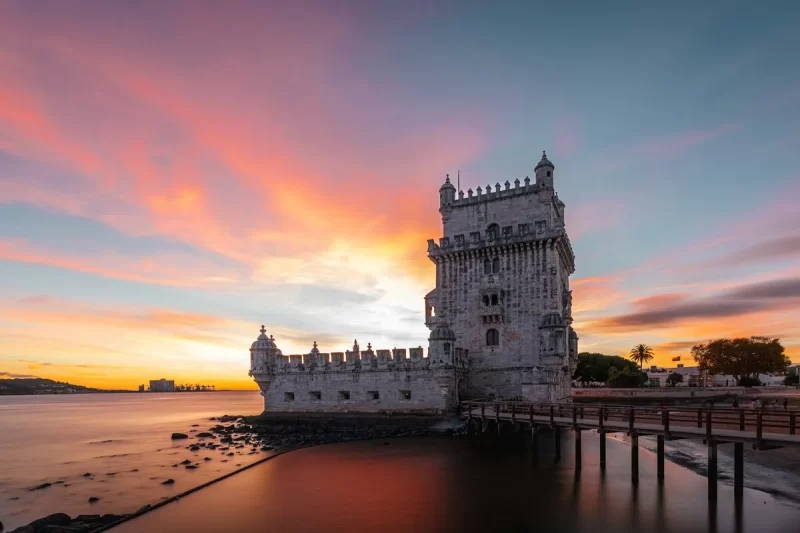 A icônica Torre de Belém em Lisboa sob um céu dramático de pôr do sol com cores vibrantes de laranja, rosa e azul, refletindo-se na água calma e destacando sua arquitetura manuelina. Torre de Belém ao Pôr do Sol: Um Espetáculo de Cores em Lisboa